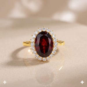May include: A gold ring featuring a large, oval-cut deep red garnet gemstone at the centre. The garnet is surrounded by a halo of small, round, clear white stones set in a scalloped gold frame. The band is a simple, polished gold metal.