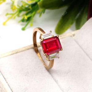 Op de afbeelding: Een gouden ring met een grote, smaragd geslepen rode edelsteen en twee kleinere, baguette geslepen heldere edelstenen aan weerszijden.