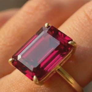 May include: A gold ring featuring a large, rectangular, emerald-cut deep red gemstone held in place by four gold prongs. The stone has a faceted surface that reflects light, and the band is a simple, smooth gold metal wire design.