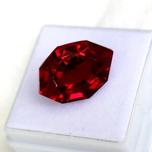 Peut inclure: Une pierre précieuse rouge foncé, taillée en forme octogonale. La gemme est posée sur une surface blanche, et les facettes reflètent la lumière, créant un aspect brillant. La pierre est d'une couleur riche et saturée.