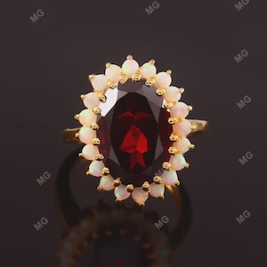 May include: A gold ring with a large, oval-cut, deep red gemstone at the centre, encircled by a halo of smaller, pear-shaped, white gemstones. The ring's band is a simple, polished gold.