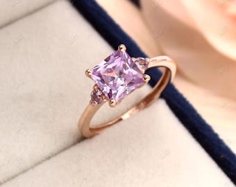 May include: A rose gold ring featuring a square, light purple gemstone as the primary focal point. Two smaller, round purple gemstones flank the main stone. The ring is set against a light-colored background.
