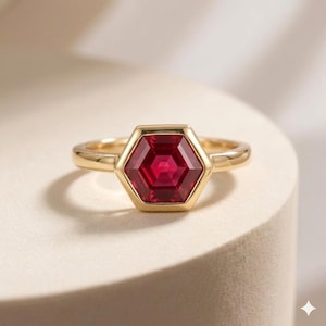 Op de afbeelding: Een gouden ring met een centrale dieprode edelsteen in een zeshoekige slijpvorm. De steen is gezet in een gladde, gepolijste gouden zetting die de zeshoekige vorm volgt, gemonteerd op een eenvoudige, slanke gouden ring met een gepolijste afwerking.