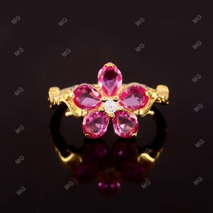 Può includere: Un anello dorato con un design a fiore con cinque petali di pietre preziose rosa e una gemma bianca centrale. La fascia dell'anello ha un design ornato. L'anello è esposto su una superficie nera riflettente.