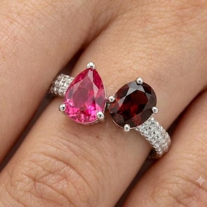May include: A silver-toned ring featuring two prominent gemstones: a bright pink pear-cut stone and a deep red oval-cut stone. The band is encrusted with small, clear, sparkling stones along the sides, creating a detailed and textured appearance.
