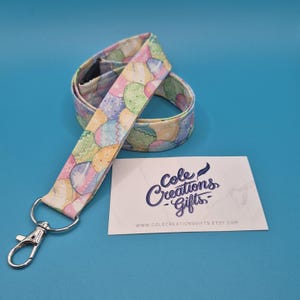 May include: A patterned lanyard featuring pastel-colored Easter eggs. It has a silver-tone clasp and a business card with the text "Cole Creations Gifts" and a website address. The background is blue.