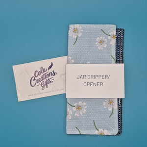 May include: A light blue fabric jar gripper and opener featuring a white floral pattern and a dark textured underside. A white paper band is wrapped around the centre with the text JAR GRIPPER/OPENER, accompanied by a white business card with the text Cole Creations Gifts.