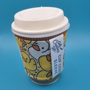May include: A disposable coffee cup with a white lid and a duck-themed cup cozy. The cozy is yellow with a pattern of white ducks and black outlines. A white tag on the cozy reads "Cup Cozy Sleeve" and "Cole Creations Gifts."