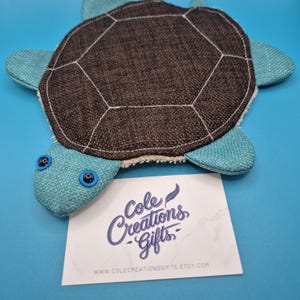 May include: A plush turtle-shaped item with a brown shell and blue flippers and head. The turtle has black and blue eyes. A white card with the text "Cole Creations Gifts" and a website address is in front of the turtle.