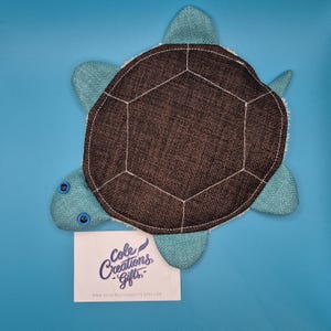 May include: A plush turtle-shaped item with a brown, geometric patterned shell and light blue flippers and head. The turtle has white stitching and blue eyes. A white card with the text "Cole Creations Gifts" is at the bottom.