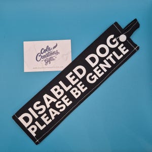 May include: Black fabric tag with white text saying "DISABLED DOG PLEASE BE GENTLE." The tag has a loop and a white button. A business card with the text "Cole Creations Gifts" is also visible.
