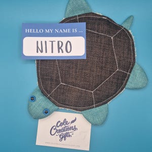 May include: A handmade turtle-shaped item with a dark gray shell and teal flippers. A blue name tag reads "HELLO MY NAME IS... NITRO". A small white card with the text "Cole Creations Gifts" is also present.