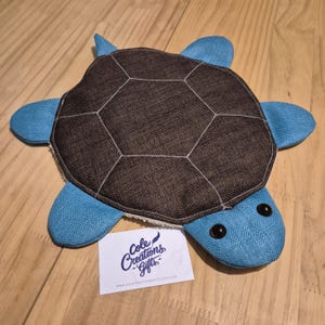 May include: A handcrafted turtle soft toy. The turtle features a brown, segmented shell with white stitching, and blue flippers, head, and tail. The eyes are black. A white card with the text "Cole Creations Gifts" is in the image.