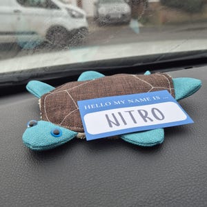 May include: A plush turtle toy with a brown shell and blue flippers and head. The toy has a name tag that reads "HELLO MY NAME IS... NITRO". The turtle is resting on a dark surface.