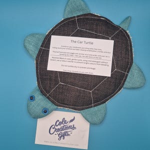 May include: A teal and gray turtle-shaped car cleaning pad. The pad has a dark gray shell and teal flippers and head. The product has a white border and includes a care instruction card. The product is from Cole Creations Gifts.