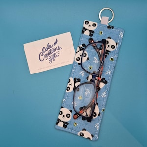 May include: A blue glasses case featuring a panda bear design, holding a pair of spectacles. The case has a key ring and a white tag with the text "Cole Creations Gifts". The spectacles have a black frame with red detailing.
