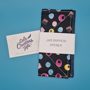 May include: A rectangular black fabric jar gripper with a colourful candy print featuring pink, blue, and yellow shapes. A white paper band across the centre reads JAR GRIPPER/OPENER, and a business card for Cole Creations Gifts is placed beside it.