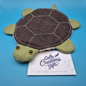 May include: A turtle-shaped fabric coaster featuring a dark brown shell with white stitched hexagonal patterns and light green limbs. The turtle has two yellow and black plastic eyes. A white rectangular card with the text Cole Creations Gifts is placed underneath the turtle.