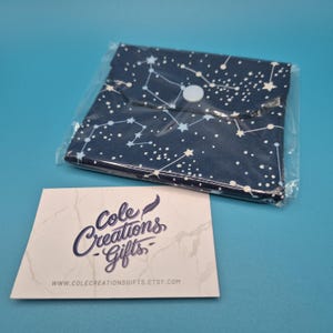May include: A dark blue fabric pouch featuring a white constellation and star pattern, secured with a single white snap button. The pouch is enclosed in clear plastic packaging and is accompanied by a white card printed with Cole Creations Gifts.