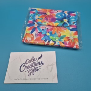 May include: A square fabric pouch featuring a vibrant, multi-coloured floral pattern in shades of pink, orange, yellow, and blue, enclosed in a clear plastic sleeve. Beside it lies a white rectangular card with the text Cole Creations Gifts.