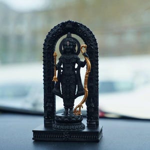 May include: A black statue of a deity, possibly Hindu, standing within an ornate archway. The figure holds a golden bow and arrow, with intricate detailing on the clothing and arch. The statue is on a decorative base.