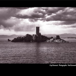 May include: A black and white photograph of a stone tower on a small island in the middle of a body of water. The sky is cloudy and there are mountains in the distance.