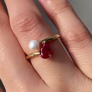 Op de afbeelding: Een gouden ring met een witte parel en een druppelvormige, dieprode edelsteen. De ring heeft een uniek, asymmetrisch ontwerp, waarbij de parel en de edelsteen aan weerszijden van de band zijn geplaatst.