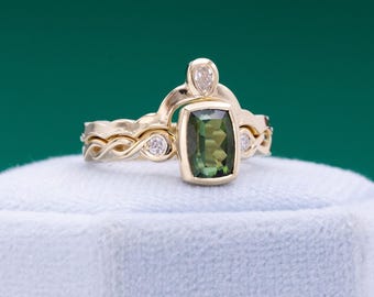 May include: A gold ring set featuring a rectangular green gemstone and a smaller pear-shaped diamond. The band has a unique, intertwined design with small accent diamonds. The ring is displayed on a white surface against a green backdrop.