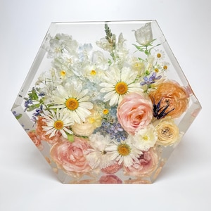 May include: A clear, hexagonal resin block encasing a vibrant floral arrangement. The flowers include white daisies with yellow centers, peach roses, and other colorful blooms. The arrangement is a decorative piece.