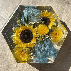 May include: A hexagonal resin block with a preserved flower arrangement. The arrangement features yellow sunflowers, yellow roses, and blue hydrangeas.