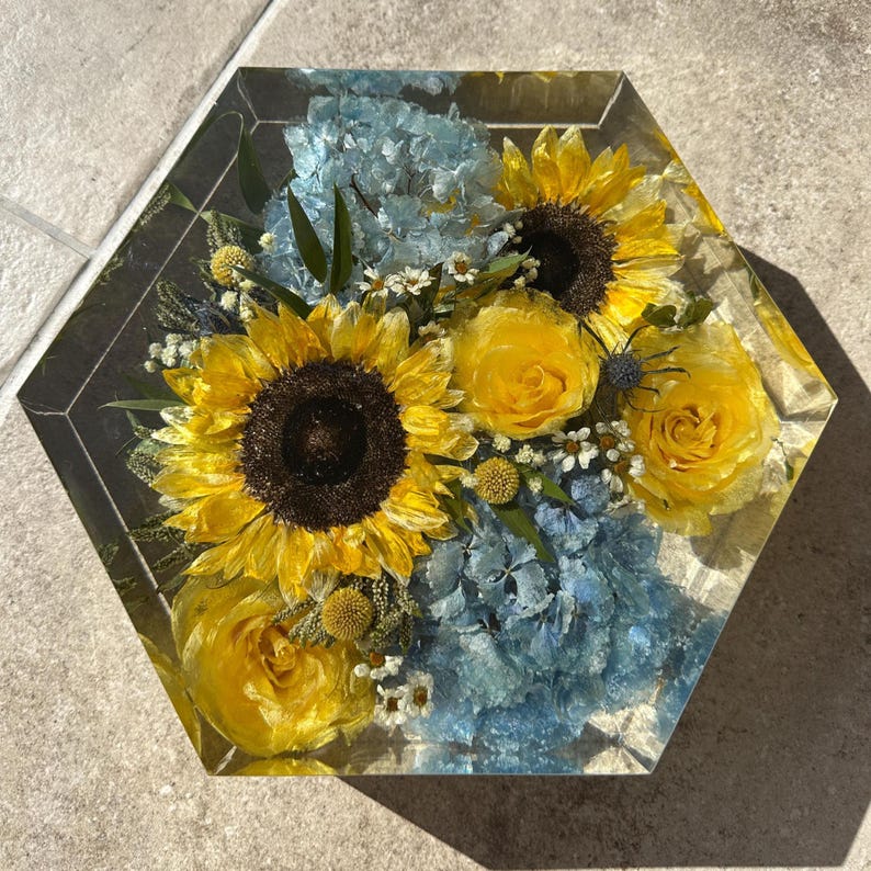 May include: A hexagonal resin block with a preserved flower arrangement. The arrangement features yellow sunflowers, yellow roses, and blue hydrangeas.