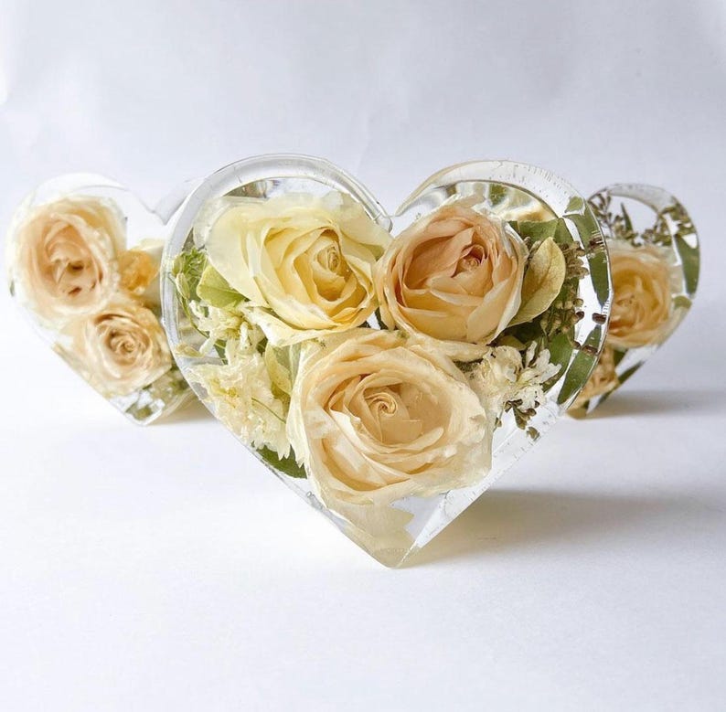 May include: Three heart-shaped resin paperweights, each encasing dried cream and peach-colored roses and greenery. The clear resin allows the flowers to be fully visible, creating a unique decorative item.