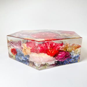 May include: A hexagonal clear resin box filled with pressed flowers in shades of pink, red, blue, and white. The flowers are arranged in a random pattern and are visible through the clear resin.