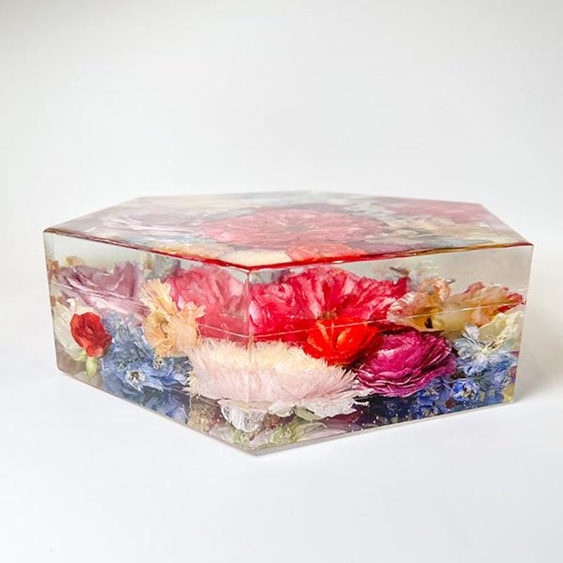 May include: A hexagonal clear resin box filled with pressed flowers in shades of pink, red, blue, and white. The flowers are arranged in a random pattern and are visible through the clear resin.