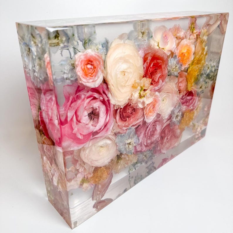 Puede incluir: Un bloque rectangular transparente lleno de flores preservadas en tonos rosa, melocot&oacute;n y blanco. Las flores est&aacute;n dispuestas en un grupo denso, creando una hermosa y delicada exhibici&oacute;n.