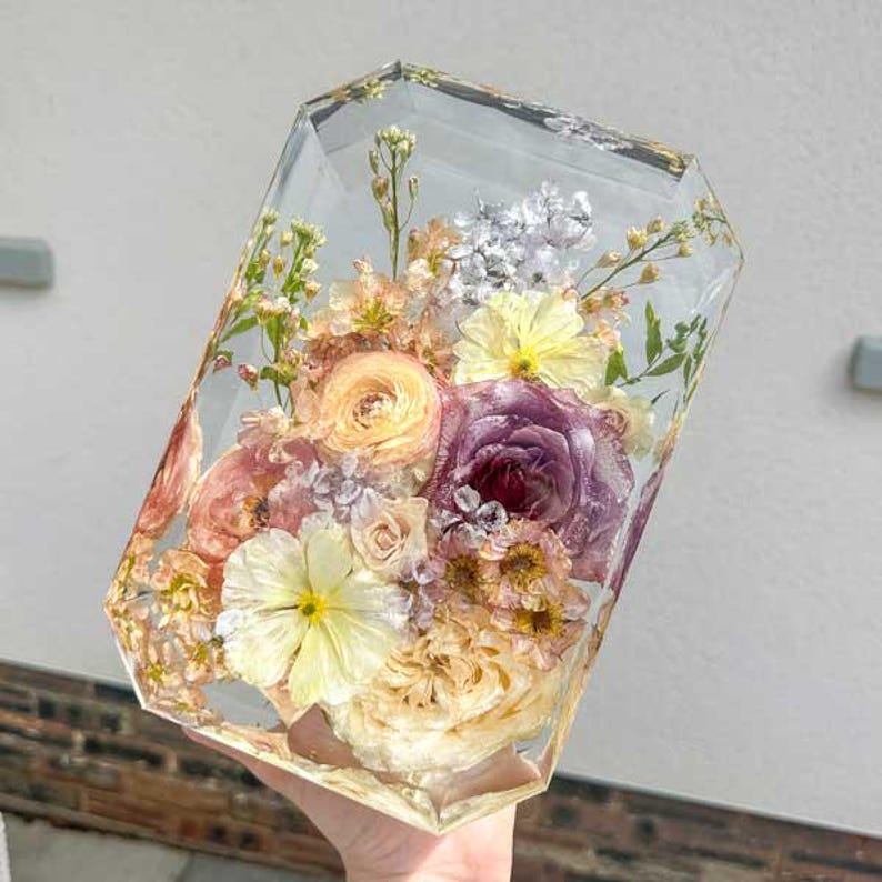 May include: A clear, geometric resin block encasing a vibrant floral arrangement. The flowers include yellow, purple, and peach-colored blooms, with hints of silver foliage. The arrangement is a decorative piece, perfect for home decor.