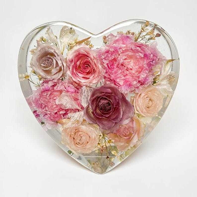 May include: A heart-shaped resin paperweight filled with an assortment of preserved flowers. The flowers are in shades of pink, rose, and cream, with some yellow accents. The clear resin allows a full view of the floral arrangement.
