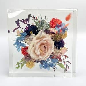 Puede incluir: Un bloque cuadrado de resina transparente con un arreglo de flores preservadas. El arreglo presenta una rosa grande de color rosa p&aacute;lido, rodeada de flores m&aacute;s peque&ntilde;as en tonos de azul, rojo y morado.