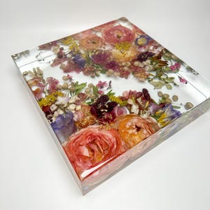 Puede incluir: Un bloque de resina cuadrado con una variedad de flores y follaje prensados en tonos de rosa, morado, amarillo y verde. Las flores est&aacute;n dispuestas en un patr&oacute;n suelto y org&aacute;nico, y la resina es transparente y brillante.