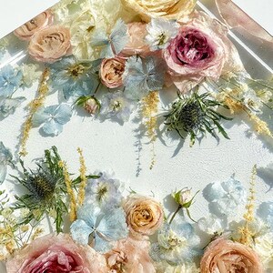 May include: A square resin art piece featuring an arrangement of dried flowers. The flowers include roses in shades of pink, peach, and cream, along with blue hydrangeas and thistle. The background is white.