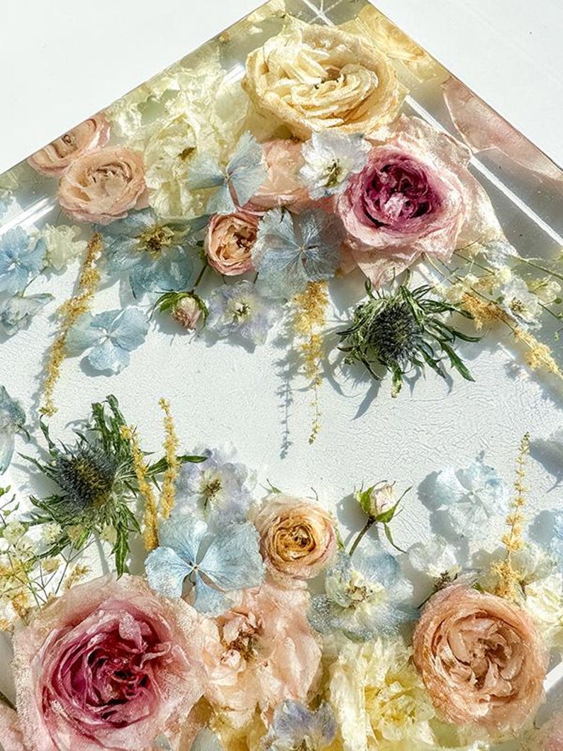 May include: A square resin art piece featuring an arrangement of dried flowers. The flowers include roses in shades of pink, peach, and cream, along with blue hydrangeas and thistle. The background is white.