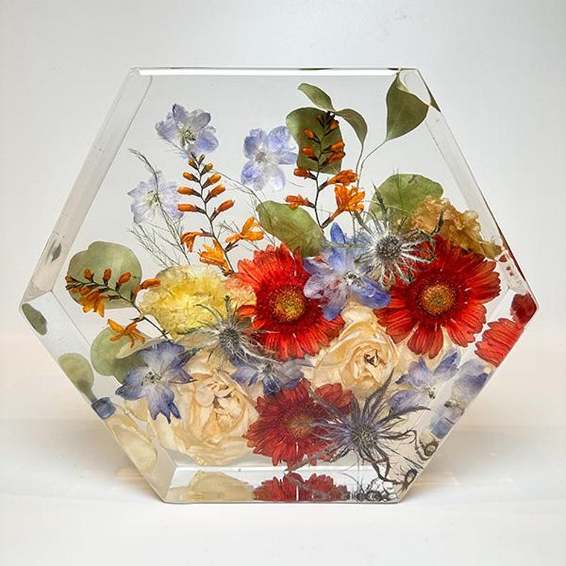 May include: A hexagonal clear resin block with a preserved flower arrangement. The arrangement features red, white, and blue flowers, including roses, carnations, and delphiniums.