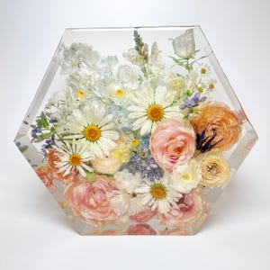 May include: A hexagonal resin block with a bouquet of preserved flowers. The flowers are a mix of white, pink, and peach, with some blue accents. The flowers are arranged in a natural, flowing style.