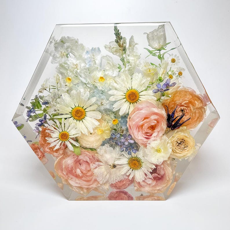 May include: A hexagonal resin block with a bouquet of preserved flowers. The flowers are a mix of white, pink, and peach, with some blue accents. The flowers are arranged in a natural, flowing style.