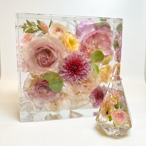 May include: A clear glass cube filled with preserved pink, yellow, and white flowers. A smaller clear glass pyramid with preserved flowers sits in front of the cube. A gold ring is displayed on top of the pyramid.