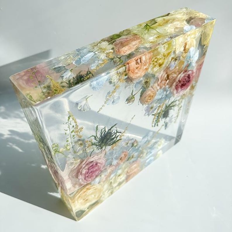 May include: A clear, angled resin block encasing a variety of dried flowers. The flowers include roses in shades of pink, peach, and cream, with accents of blue and green foliage. The block is transparent, allowing the flowers to be fully visible.