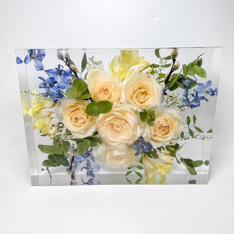 May include: A clear rectangular resin block encasing a floral arrangement. The arrangement features cream-colored roses, blue delphiniums, yellow freesia, and green foliage. The flowers are arranged in a visually appealing composition, creating a decorative piece.