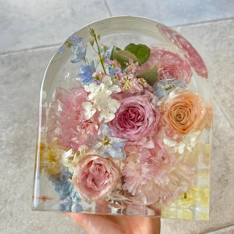 May include: A clear, arched resin block encasing a vibrant floral arrangement. The bouquet features pink, peach, and white roses, carnations, and delicate blue and white blossoms. The flowers are preserved within the transparent resin, creating a decorative display.