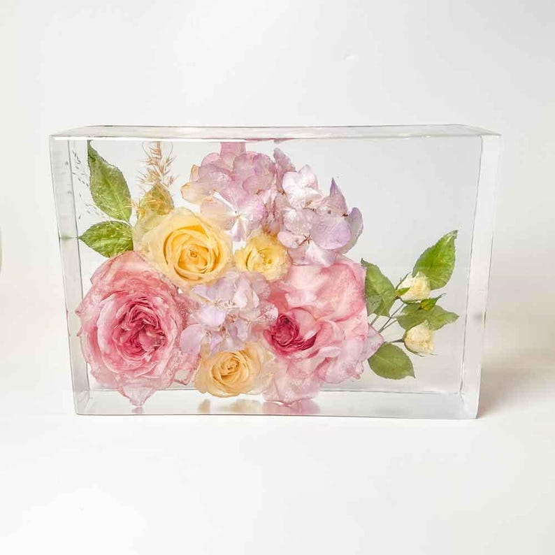 Puede incluir: Una caja rectangular transparente con un ramo de flores preservadas. Las flores son rosas, amarillas y moradas. Las flores est&aacute;n dispuestas en un grupo en el centro de la caja.