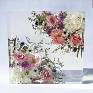 May include: A square clear acrylic block with pressed pink, white, and purple flowers inside. The flowers are arranged in a bouquet-like fashion.